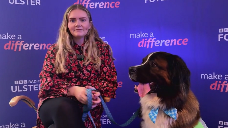 A blonde woman sits on a chair holding a dog lead with a dog on the end