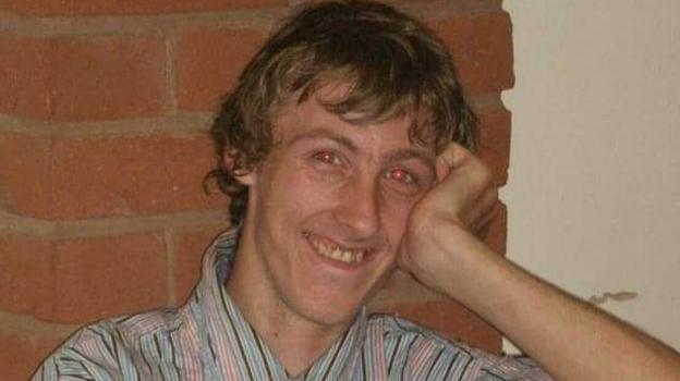 A man with messy, dark blonde/light brown hair is looking towards the camera and smiling
