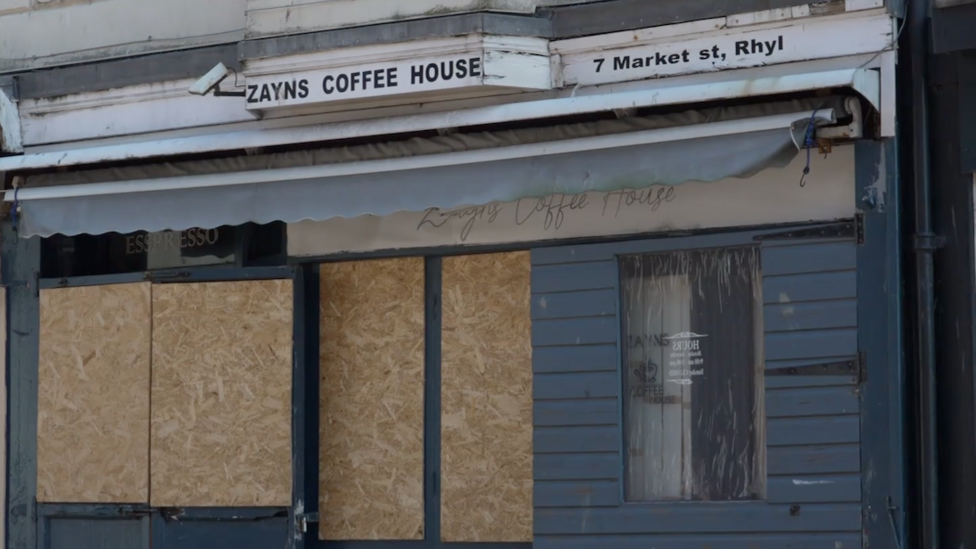 Boarded up cafe front with the word's Zayns Coffee House above a blue shut canopy. One of the blue window frames and doorway are boarded with plain plywood
