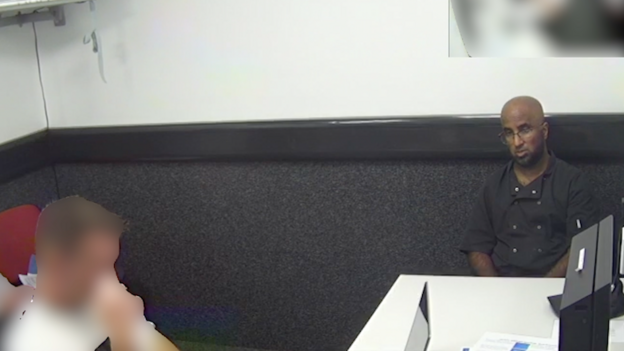 Image shows a blurred out police officer in a room opposite Mustafa Iqbal who is wearing a black catering-style shirt, bald and wearing glasses. A white table is between him and the officer.