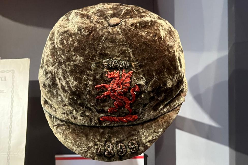 An old sports cap which looks like a velvet material. It's a green/ brown colour with a faded embroidered red dragon and the date 1899 on the brim.