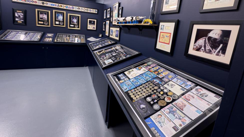 A display case containing space flight memorabilia and artefacts. On the walls around the case are framed photos of astronauts and artefacts.