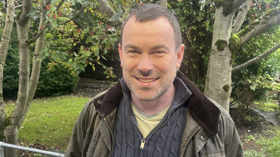 Dave Neil is outdoors. He has close cropped hair, stubble and is wearing a green outdoor jacket over a grey top and a yellow T-shirt. There is grass and a trees behind him.