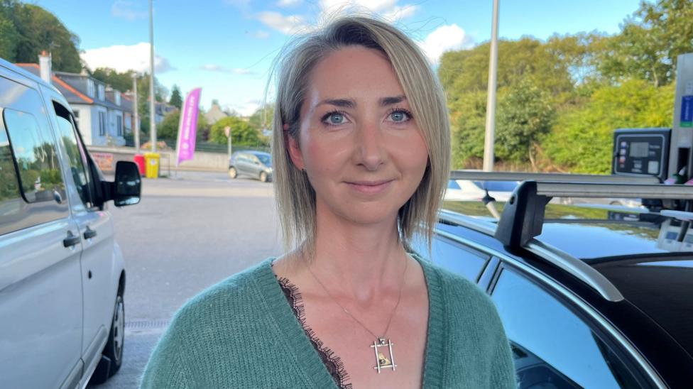 Crofter and musician Amy Henderson is standing in a petrol station forecourt. She has short blonde hair and is wearing a green cardigan. There is a car and also a van behind her.