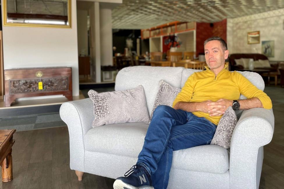 James Dunbar is sitting on a sofa in a warehouse. He has short hair and is wearing a yellow fleece and blue jeans.