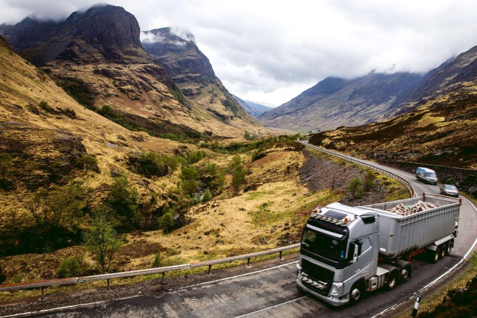 A lorry comprising a silver-coloured tractor unit hauling a silver-coloured trailer on a winding stretch of the A82 through Glen Coe.