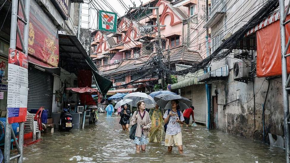 People holding umbrellas and walking down a narrow flooded street in Bangkok