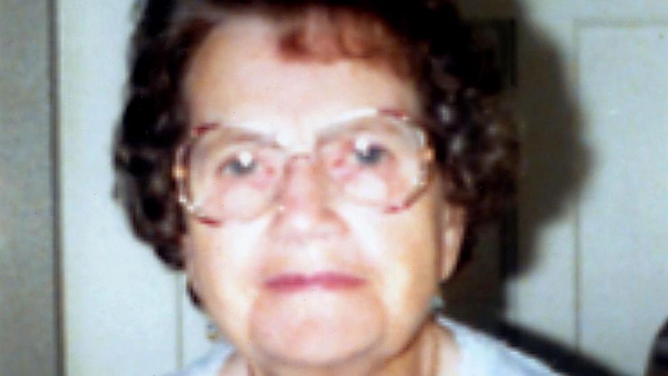 Ethel Hall - Portrait photograph of an older person wearing large glasses and a light patterned top with a pendant necklace, seated indoors against a plain background.