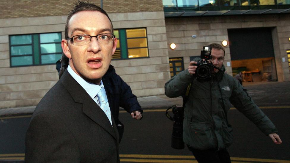 Colin Norris outside Newcastle Crown Court in 2008 - A person in a suit and glasses walks outside a building while a photographer points a camera with flash attachment towards them on a city street.