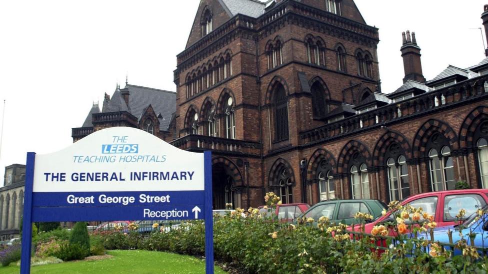 Exterior view of Leeds General Infirmary, showing the hospital's historic red-brick building behind a large sign reading "The Leeds Teaching Hospitals - The General Infirmary, Great George Street", with parked cars and flowerbeds in the foreground.