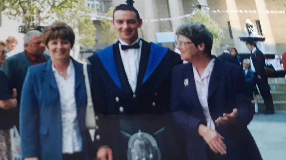 Colin Norris - A graduate in a black gown with blue trim stands outdoors between two women in jackets, posing for a photograph at a graduation ceremony. Other attendees and a building with columns are visible in the background.