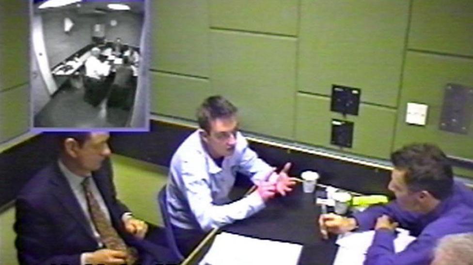 CCTV-style image showing three people seated around a table in a police interview room, with papers and cups on the table and an inset image in the corner showing a wider view of the room.