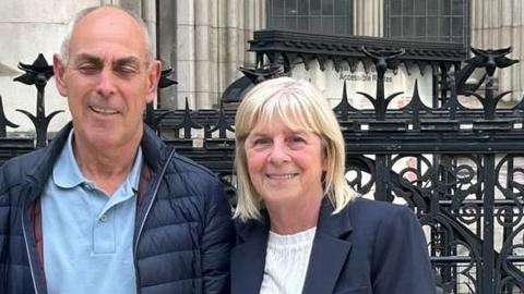 June and Raymond, Colin's mum and stepdad - Two people stand side by side outside a court building, posing for a photograph in front of ornate black railings and stone columns.