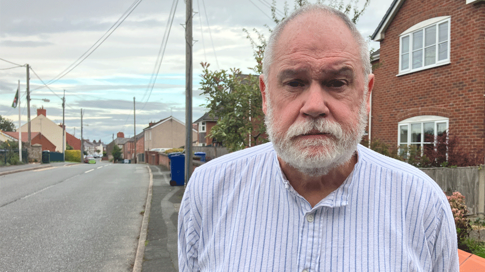 Mark Andrew is wearing a white shirt with blue stripes. He is standing on the pavement of street of red brick houses