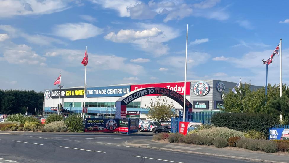 A large Trade Centre UK car dealership is pictured beside a road, with a prominent entrance arch reading “Welcome to Trade Centre UK”. The modern showroom building features large advertising banners, including signs promoting cars from £99 a month and “Drive Away Today”.