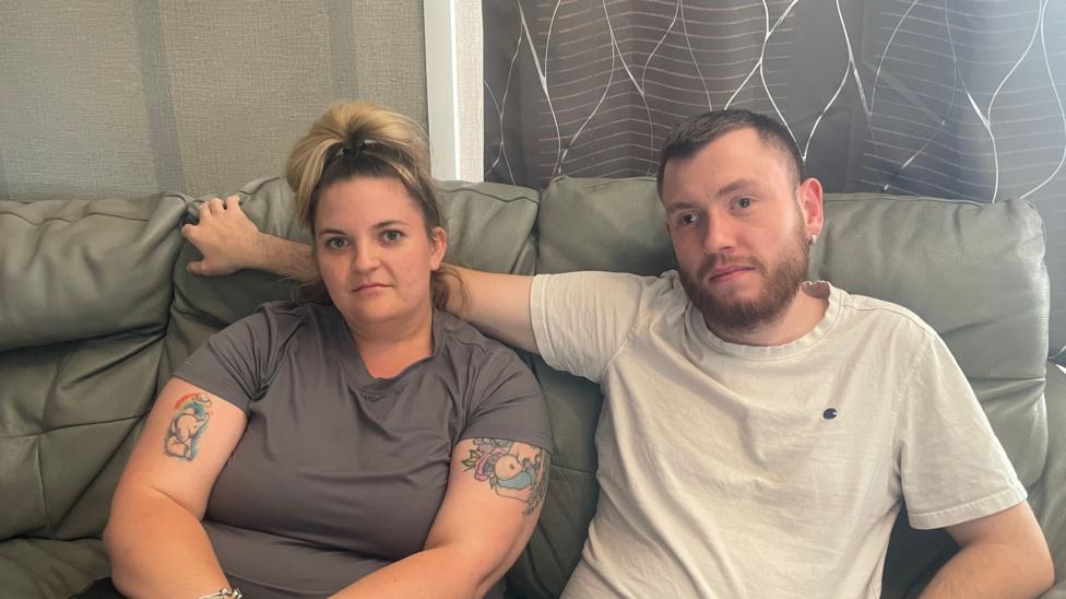 A couple are seated together on a grey leather sofa in a living room. One person is wearing a grey T-shirt and has visible tattoos on both upper arms, while the other is wearing a white T-shirt and has an arm resting along the back of the sofa behind the other person. Both are looking towards the camera.