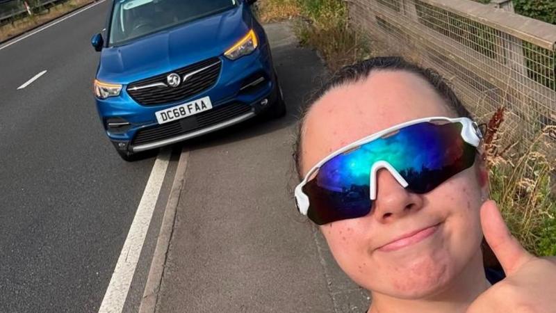 A woman stands at the side of a dual carriageway, with a thumbs up to the camera in this selfie photo. She is wearing a navy vest top and reflective blue sunglasses. Behind, parked half on the kerb, is a blue Vauxhall car with its orange hazard lights on.