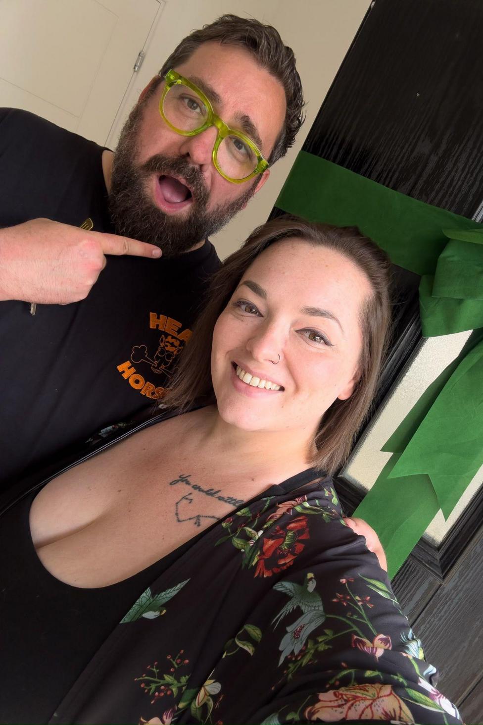 A couple stand in front of a front door with a green silk bow tied onto it. The door is open and they are stood inside the house. A woman stands on the right and is grinning, she has a black floral kimono on over a black scoop neck t-shirt. She has straight brown hair. On her left is her husband who is grinning with his mouth open, and pointing to the door. He is wearing luminous yellow framed glasses. He has dark brown hair and facial hair and is wearing a black t-shirt. 