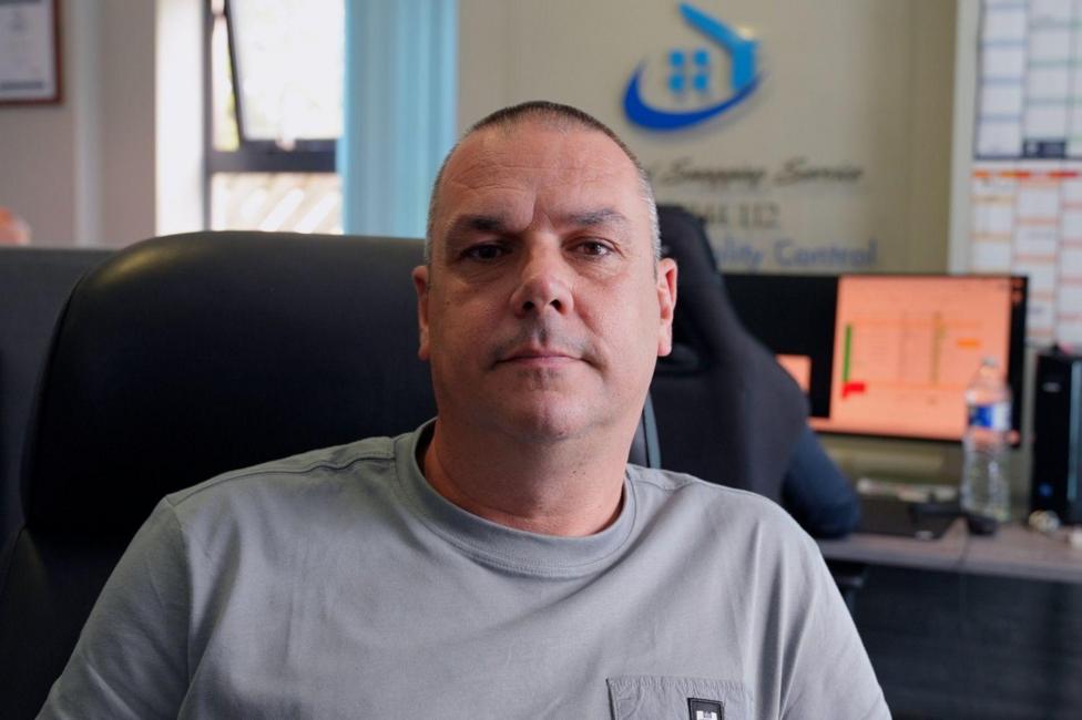 A man smiles at the camera, he has short cropped hair and is wearing a grey t-shirt. He is sat in an office. 