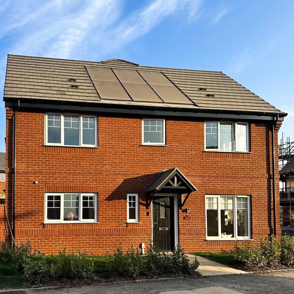 A new build home, with red bricks and five windows. There is a black wooden door in the middle and there is a grass section in the front. There are bushes along the front. There are solar panels on the roof and grey roof panels. 