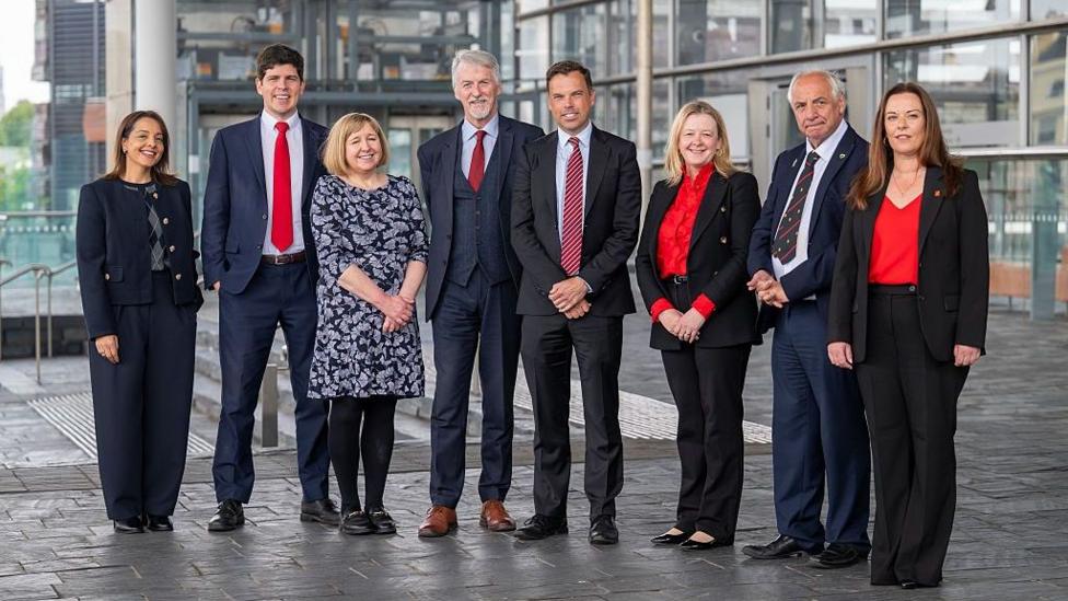The picture shows eight of Welsh Labour's nine MSs posing for a photograph outside the Senedd. They are smiling and looking at the camera.