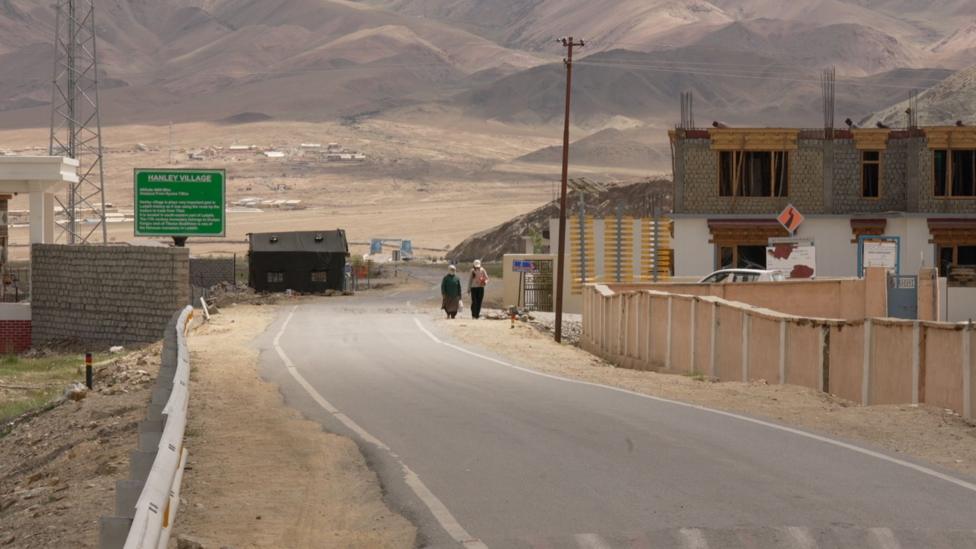 Two women seen walking at a distance on a road in Hanle. There is construction work happening on the side as well.