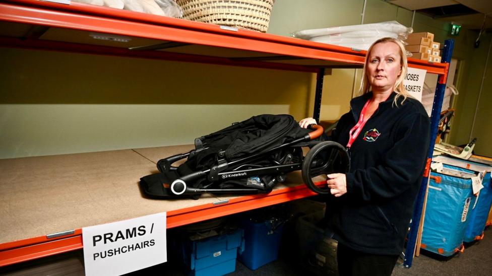 A woman with shoulder length blonde hair, wearing a dark fleece and trousers stands in a warehouse with large shelves next to her. One is labelled "Prams and Pushchairs" and has a black folded up pram on it and the woman is holding the handle.