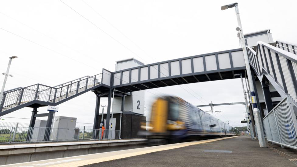 A view from a platform at the new Balgray station. The camera is on the platform ground looking upwards. A train is speeding through, but at such speed that it is blurry in the image. The stairs and bridge that connect the platforms can be seen, and a man in orange overalls watches the train go through.