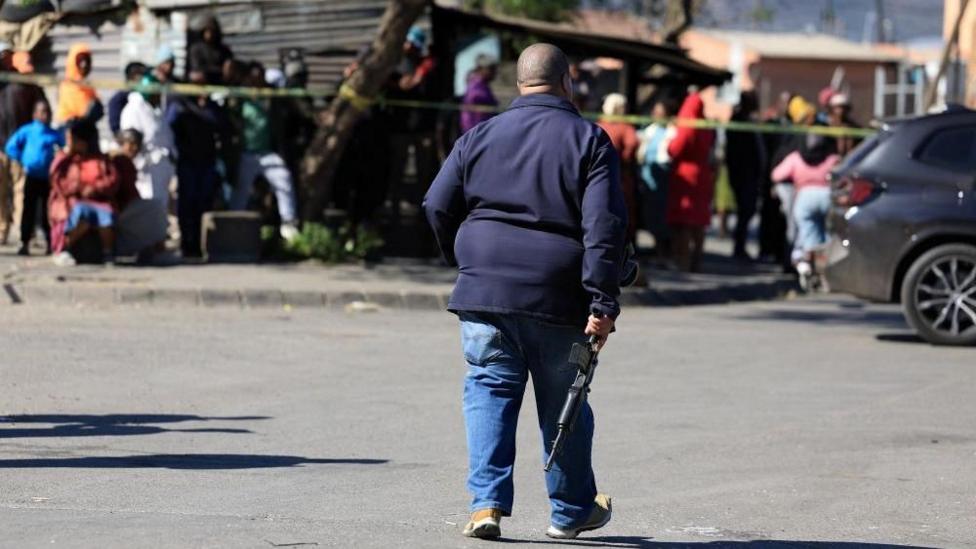 An armed guard walking down a road with people looking on from behind police tape