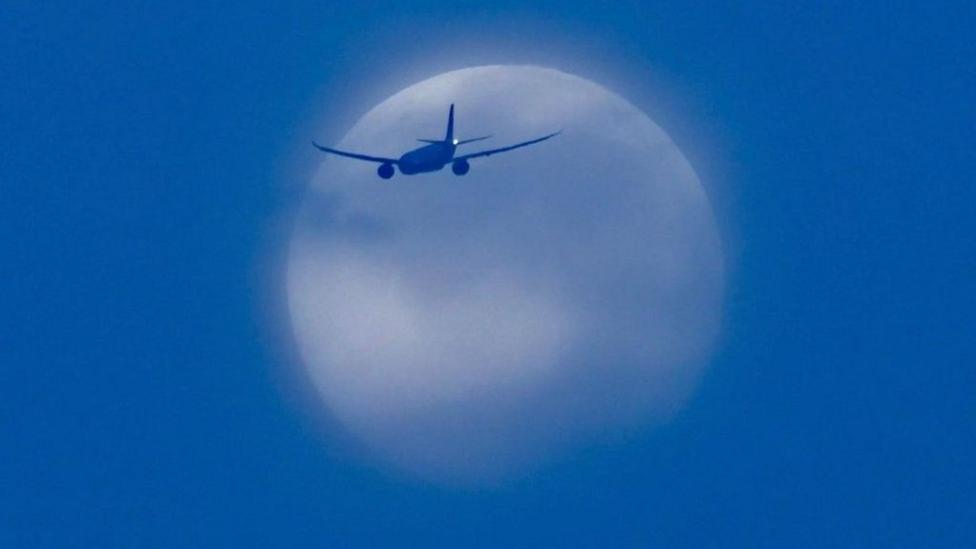 The white Moon is partially covered in blue clouds with a plane captured in front of it. It's all set in a deep blue sky