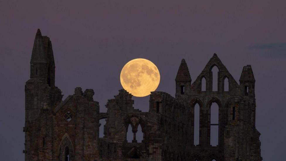 The Moon is a golden ball rising over the centre of ruins silhouetted in a grey, purple sky