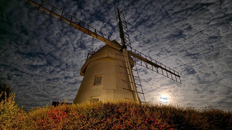 A windmill sits against a sky full of broken white clouds with the Moon glowing as it just starts to rise to the right