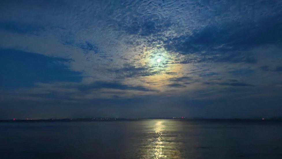 The Moon is small and white in a very blue night sky with a streak of orange reflected in the sea below