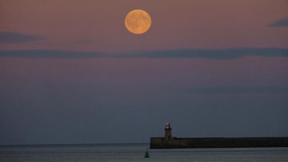 The pink sky is crossed horizontally with blue lines of cloud with a glowing orange Moon in the centre. Below a jetty with a small lighthouse juts out into the sea