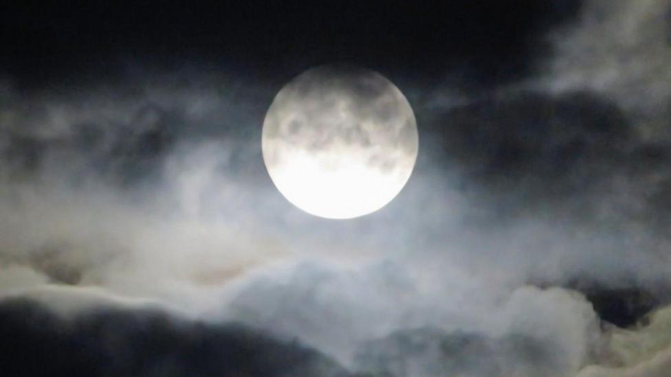 Black and grey clouds fill the sky with a bright white Moon, covered in wisps of cloud shines in the centre