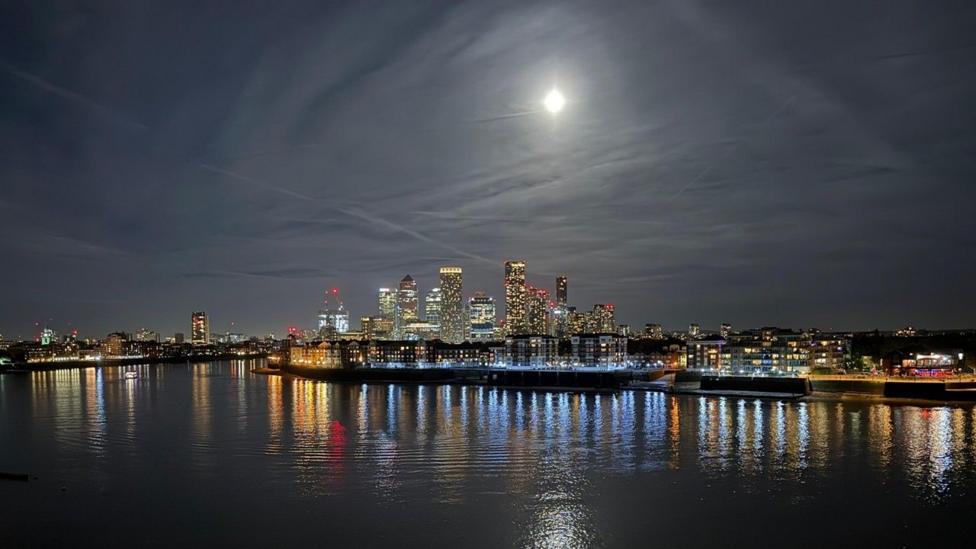 The Moon is much smaller above a wide short of buildngs lining the Thames. Mulit-coloured lights from the ground level are reflected back from the water