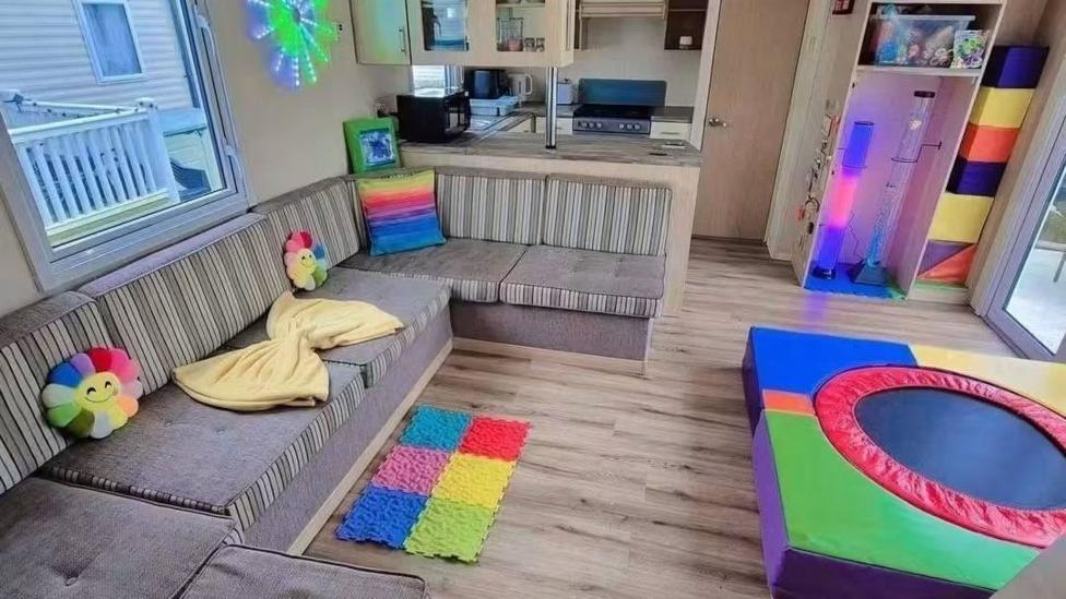 Inside of the Sen-Den caravan which shows a grey sofa with rainbow sunshine cushions and a colourful round trampoline near the sofa. 