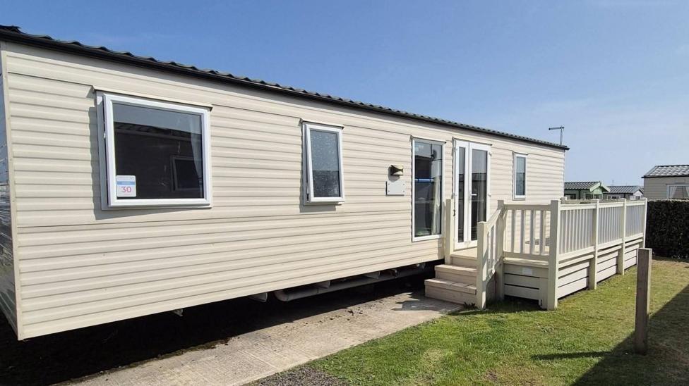 Outside the Sen-Den caravan which has a flat greyish roof and white walls with white framed windows and door. 