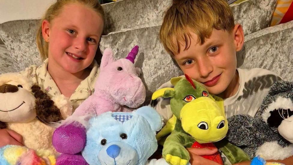Niaomi's daughter (left) and son (right) sitting on a sofa together with teddy bears in front of them. They both have blonde hair and blue eyes.