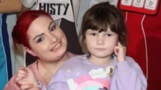 Laura (left) and her daughter Carys (right) next to each other. Laura has red hair and is wearing a pink shirt and Carys is wearing a purple shirt that has a rainbow on and has brown hair. 