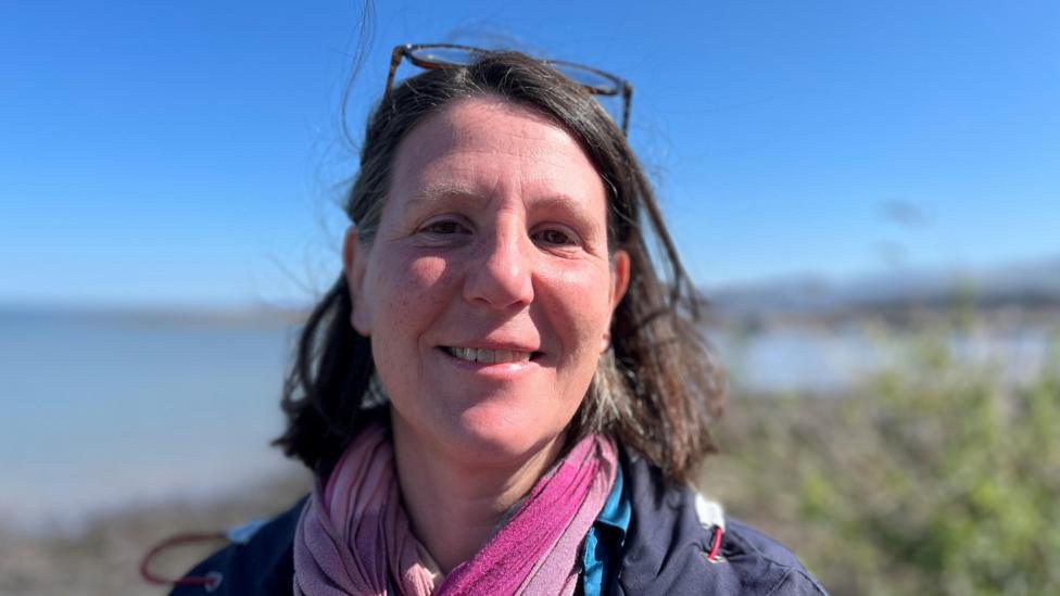 Dr Linda Osti, smiling at the camera with a coastal view behind her. She has sunglasses on her head and is wearing a blue coat with a pink scarf.