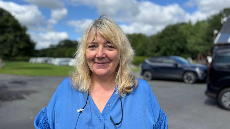 Bethan, who has long blonde hair and is wearing a blue blouse. She is smiling and looking at the camera with a car park and green field with caravans on it behind her.