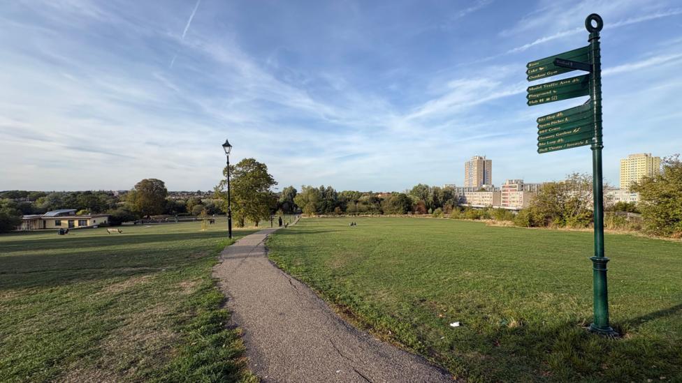 A wide, open park landscape with a paved footpath running through a large grassy area under a bright blue sky streaked with thin clouds and aircraft contrails. A green directional signpost stands on the right side of the image, while a traditional lamppost lines the path. Trees border the park, and several high-rise residential buildings can be seen in the distance.
