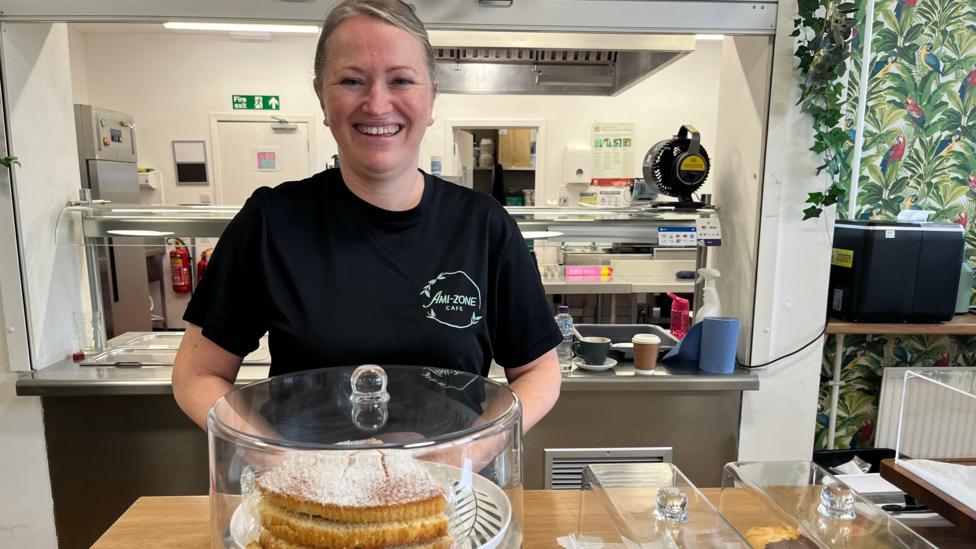 A woman in a black t-shirt, standing behind a Victoria Sponge cake in a cafe
