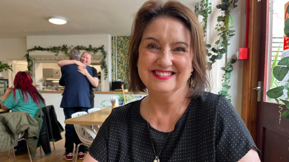 A woman with chin-length dark hair. She is smiling and wearing pink lipstick and a black top. She is sitting a cafe and a woman can be seen giving a man a hug in the background.