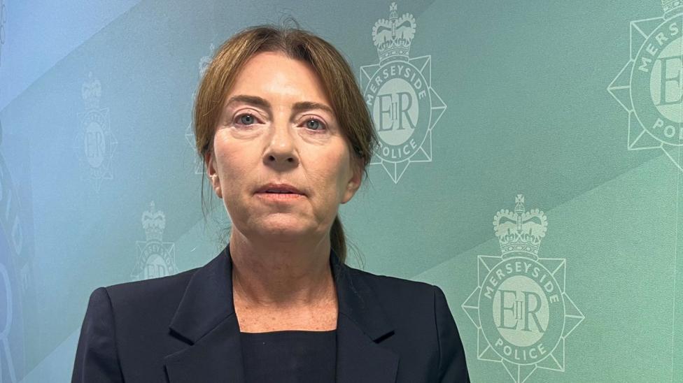 Det Supt Rachel Wilson with long brown hair tied back wearing a black top and black blazer standing in front of a green wall with the Merseyside Police logo on it.