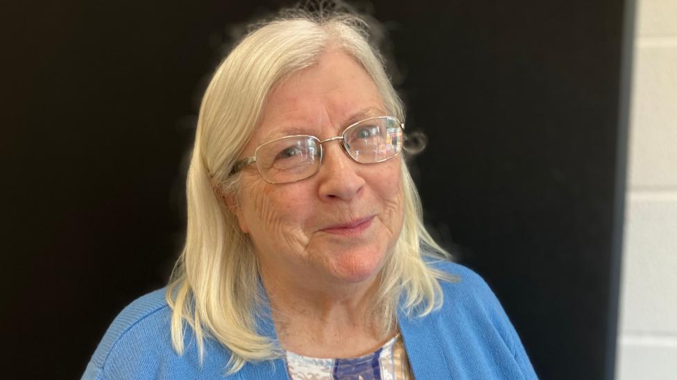 A woman in glasses with shoulder length blonde hair. She is wearing a blue cardigan and is stood against a black background 