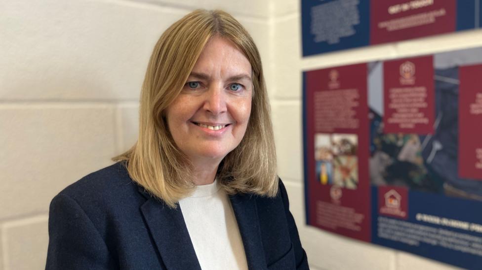A woman with shoulder length hair, wearing a blue jacket over a cream top. She is smiling at the camera, and there are posters to the side of her