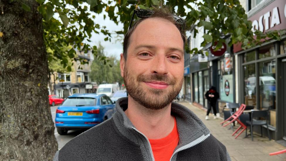 A man with a beard wears a grey fleece and orange top.