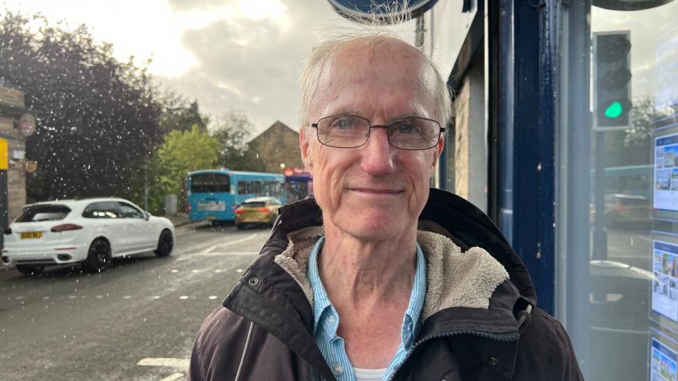 A man with white hair wears a raincoat and blue shirt.